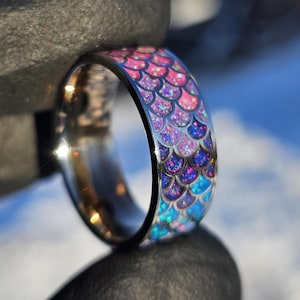 May include: A close-up of a silver ring with a mermaid scale design. The scales transition from pink to purple to blue, with glitter accents. The ring is held against a dark gray background, with a blurred blue sky in the background.