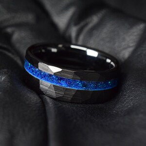 Puede incluir: Un anillo de carburo de tungsteno negro con una incrustaci&oacute;n de purpurina azul. El anillo tiene un acabado cepillado y un borde biselado.
