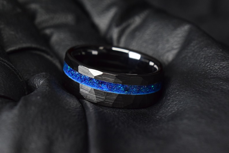 Puede incluir: Un anillo de carburo de tungsteno negro con una incrustaci&oacute;n de purpurina azul. El anillo tiene un acabado cepillado y un borde biselado.