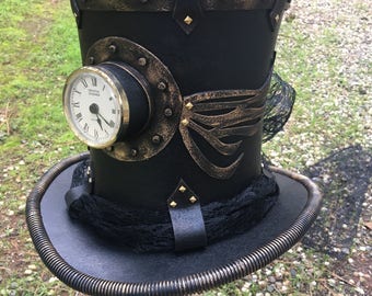 May include: A black top hat with gold accents and a clock face on the side. The hat has a black and gold band around the base and a black lace trim.