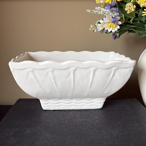 Könnte beinhalten: Eine weiße, rechteckige Keramikschale mit gewelltem Rand und strukturierten Seiten. Die Schale steht auf einer dunklen Oberfläche, mit einem Blumenarrangement und einer dekorativen Wandverkleidung im Hintergrund.