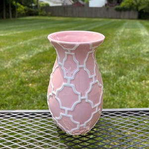 Peut inclure: Un vase en céramique rose clair orné d'un motif géométrique en relief blanc en forme de quatrefeuille sur toute sa surface. Le vase présente un col évasé et un corps arrondi, avec une finition lisse et brillante contrastant avec le motif.