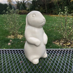 Könnte beinhalten: Eine weiße Keramik-Hasenfigur mit gesprenkeltem Finish. Der Hase steht aufrecht mit hochgestellten Ohren und zusammengefügten Pfoten.