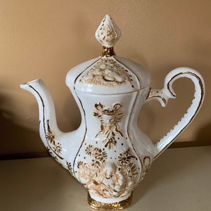 May include: White ceramic teapot with gold accents and floral designs. The teapot has a lid with a pointed top and a handle on the side.