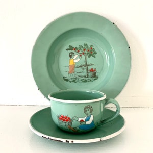Puede incluir: Un juego de té vintage para niños de esmalte verde con un platillo, una taza y un plato. La taza y el plato muestran a un niño recogiendo manzanas de un árbol, mientras que el platillo muestra a un niño sosteniendo una cesta de manzanas.
