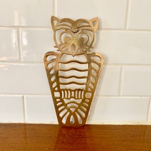 Puede incluir: Una decoración de pared de metal dorado en forma de búho con un diseño recortado. El búho tiene un cuerpo redondo con una cola puntiaguda y ojos grandes y redondos.