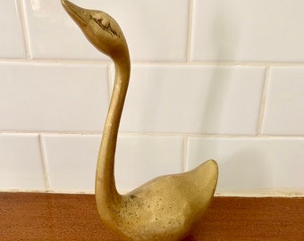 Può includere: Una statuetta di cigno in metallo dorato con un collo lungo e una testa dettagliata. Il cigno è in piedi su una superficie di legno.
