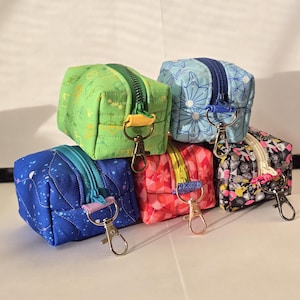 May include: A stack of six small, square fabric pouches with zippers and metal clasps. The pouches are in various colors, including green, blue, red, and black, each with a unique pattern. The zippers and clasps are in coordinating colors.