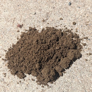 May include: A pile of dark brown, granular material, possibly soil or a similar substance, sits on a rough, light gray concrete surface. Small, dark pebbles and reddish-brown fragments are scattered around the pile.