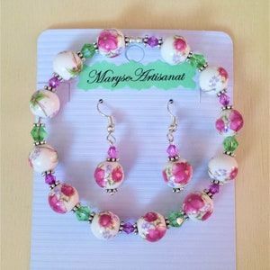 May include: A white and pink floral bracelet and matching earrings. The bracelet is made of white ceramic beads with pink floral designs and green and purple crystal accents. The earrings are silver with pink floral designs and purple crystal accents. The jewellery is displayed on a light blue card with the text "Maryse Artisanat".