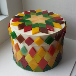 Peut inclure: Une boîte ronde blanche avec un motif en mosaïque sur le dessus et les côtés. La mosaïque est faite de petits carrés colorés dans des tons de rouge, vert, jaune et marron.