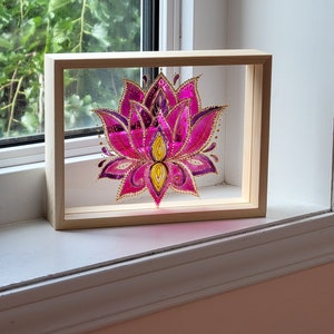 Peut inclure: Une peinture sur verre encadrée d'un lotus rose avec des accents dorés. La peinture est dans un cadre en bois et est posée sur un rebord de fenêtre.