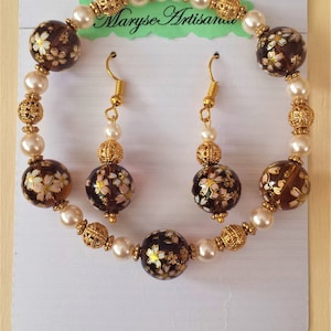 Peut inclure: Un ensemble de bijoux brun et or avec des perles de perles blanches et des motifs floraux. L'ensemble comprend un bracelet et une paire de boucles d'oreilles. Le bracelet a un fermoir en or et les boucles d'oreilles ont des crochets en or. Les bijoux sont présentés sur une carte blanche avec le texte "Maryse Artisan".