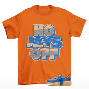 May include: An orange short-sleeve t-shirt featuring the text NO JAYS OFF in blue and tan block lettering. Below the text, a blue and orange athletic sneaker with mesh detailing and a thick sole is positioned on the lower right side of the shirt.