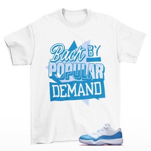 Könnte beinhalten: Weißes T-Shirt mit dem Text "Back by Popular Demand" in Blau. Unter dem Shirt ist ein Paar weiße und hellblaue Turnschuhe abgebildet. Das Shirt hat kurze Ärmel und einen Rundhalsausschnitt.