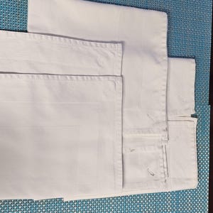 May include: White linen napkins and placemats are neatly stacked on a blue woven placemat. The napkins have a subtle grid pattern and are folded, showing their crisp edges. The placemats are also white, complementing the napkins.