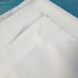 May include: Two white tablecloths with a subtle texture, folded in a corner, showing a single stitch detail.