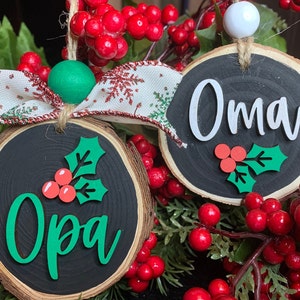May include: Two round wood slice ornaments with black paint and white lettering. The ornaments are decorated with green holly leaves and red berries. One ornament says "Oma" and the other says " Opa".