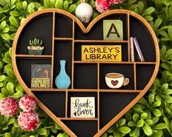 May include: Wooden heart-shaped shelf ornament with a black interior and various miniature items. Includes a small cactus, a blue vase, a book, a coffee cup, and the text "Ashley's Library" and "book lover". The ornament is surrounded by green foliage and red berries.