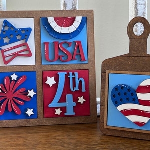 Puede incluir: Un letrero de madera con un tema de bandera estadounidense rojo, blanco y azul. El letrero está dividido en cuatro cuadrados con diferentes imágenes: una estrella, la palabra "USA", un fuego artificial y el número "4th". Una tabla de cortar de madera separada con un fondo azul y un corazón rojo, blanco y azul con estrellas.