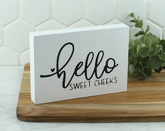 May include: White wooden sign with black text that reads "hello sweet cheeks".