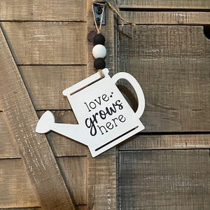 May include: White wooden watering can-shaped sign with the text "love grows here" in black script. The sign hangs from a jute string with black and white wooden beads. The background is a rustic wooden door.