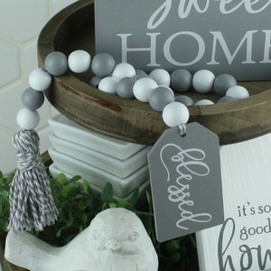 May include: A wooden bead garland with gray and white beads, a gray tassel, and a decorative tag that says "Blessed". The garland is displayed with other home decor items, including a wooden tray and signs that say "sweet HOME" and "it's so good to be home".
