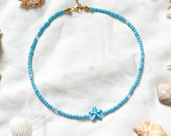 Peut inclure: Un collier de perles bleu clair avec un pendentif étoile bleu clair. Le collier est sur un fond blanc avec des coquillages.