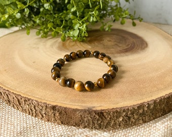 May include: A brown tiger's eye beaded bracelet with gold accents. The bracelet is on a wooden surface.