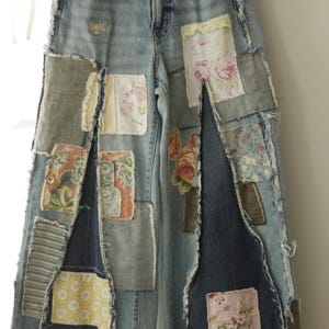 May include: Light blue denim jeans featuring a wide-leg silhouette and a patchwork design. The pants are embellished with various rectangular fabric patches in floral, striped, and geometric patterns, all with frayed edges and visible stitching.