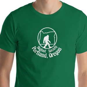 Puede incluir: Una camiseta verde con un gráfico blanco de un Bigfoot caminando dentro del contorno del estado de Oregón. El gráfico también incluye el texto "Bigfoot Society Portland, Oregon".