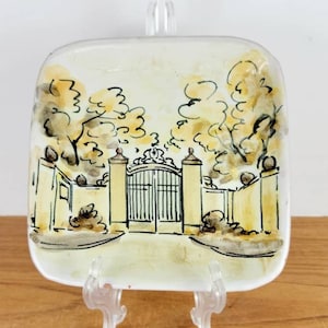 May include: A square, off-white decorative plate with a hand-painted illustration of a gate, trees, and a building. The artwork features black and yellow accents. The plate is displayed on a clear plastic stand.