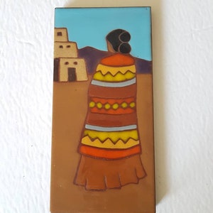 May include: Rectangular ceramic tile featuring a Southwestern design. The artwork depicts a figure in a colorful blanket, standing before a pueblo and mountains under a blue sky. The tile has a brown border and background.
