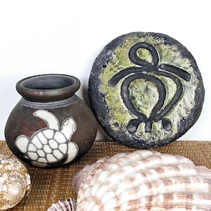 May include: A brown ceramic pot with a white turtle design, a round stone with a black turtle symbol, and a large seashell. The pot and stone are on a woven surface, suggesting a coastal or nautical theme.