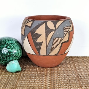May include: A terracotta-colored, round pottery bowl with a geometric design in shades of beige, gray, and rust. The bowl has a wide opening and a rounded base. A green malachite sphere and a turquoise stone are next to the bowl.