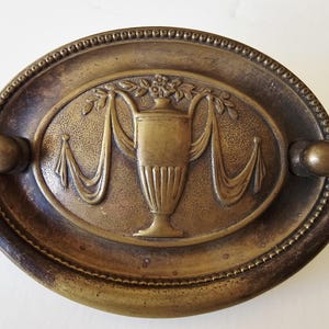 May include: Antique brass drawer pull with an oval backplate. The center features a detailed relief of a vase with draped fabric and floral accents. The pull has a decorative beaded border and curved handles.