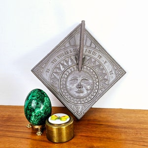 May include: A gray metal sundial with Roman numerals and a sun face design. The sundial is on a wooden surface with a green malachite egg and a brass trinket box with a butterfly design.