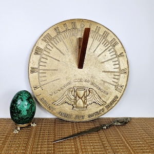 May include: A gold-toned metal sundial with Roman numerals and the inscription "I count none but sunny hours". The sundial is on a brown woven surface with a green egg and a silver letter opener.