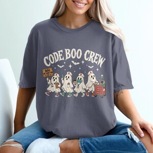 May include: A charcoal gray t-shirt featuring a graphic of four ghosts dressed as medical professionals. The text reads CODE BOO CREW and BOO WE SAVE YOU. The ghosts wear nurse caps, stethoscopes, and masks, with one pushing a medical cart.