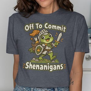 May include: A dark heather grey t-shirt featuring a cartoon frog dressed as a knight with a helmet, shield, and sword. The graphic includes the text "Off To Commit SHENANIGANS" in a light yellow font, with the frog carrying a brown backpack.
