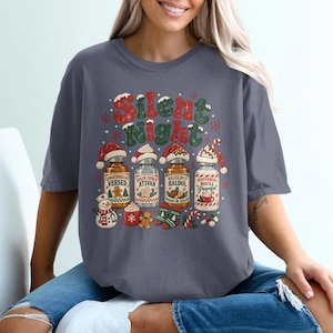 May include: A charcoal gray t-shirt featuring a festive graphic with the text Silent Night. The design includes four medicine bottles labeled Gingerbread Versed, Sugar Cookie Ativan, Hazelnut Haldol, and Peppermint Mocha Propofol, with holiday accents.
