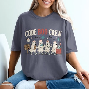 May include: A charcoal gray t-shirt featuring a graphic of four ghosts dressed as medical staff. The text reads CODE BOO CREW. The ghosts are depicted with stethoscopes, clipboards, and a medical cart, accompanied by bats and a sign that says BOO WE SHIVE YOU.