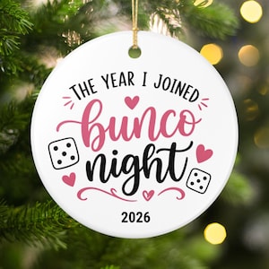 May include: A round white ceramic ornament featuring the black and pink text THE YEAR I JOINED bunco night 2026. The design includes two black and white dice icons, three small pink hearts, and decorative flourishes, suspended by a gold string.