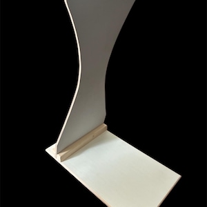 May include: A light-colored wooden display stand with a curved back and a rectangular base. The stand is made of unfinished wood and has a simple, modern design. The stand is suitable for displaying items.