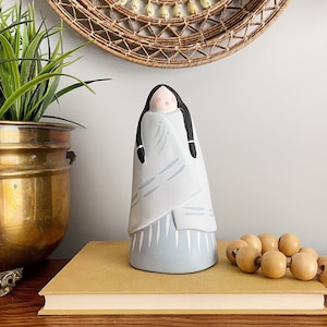 May include: A ceramic figurine of a woman wearing a blue and white patterned dress and a long black braid. The figurine is standing on a yellow book with a wooden surface behind it.