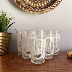 May include: Set of six vintage clear drinking glasses. Each glass features a white oval design with wheat stalks. The glasses are displayed on a wooden surface, with a woven wall hanging and a potted plant in the background.
