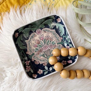 May include: Square ceramic dish with a dark blue background and a white rim. The dish features a central floral design in pink, green, and cream colors. A wooden bead necklace rests on the white fur surface next to the dish.
