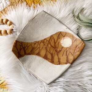 May include: A square ceramic dish with rounded corners featuring a hand-painted design of bare brown trees against a white background. A circular white moon is visible within the brown tree-filled area, which spans across the center of the dish.
