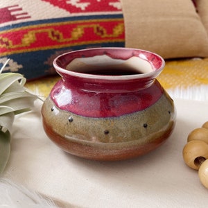 May include: A small, hand-crafted ceramic pot with a red, brown, and gray glaze. The pot has a rounded shape and a wide opening.