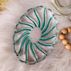 May include: Clear glass serving dish with a spiral design. The dish features teal green accents that follow the curves of the design. The dish is oval-shaped and sits on a white, fluffy surface.